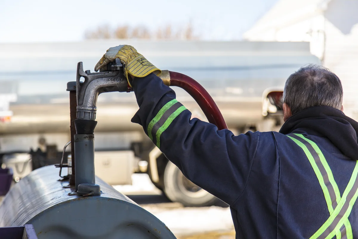 Rear view of a driver holding a hose nozzle at an outdoor oil tank connection.